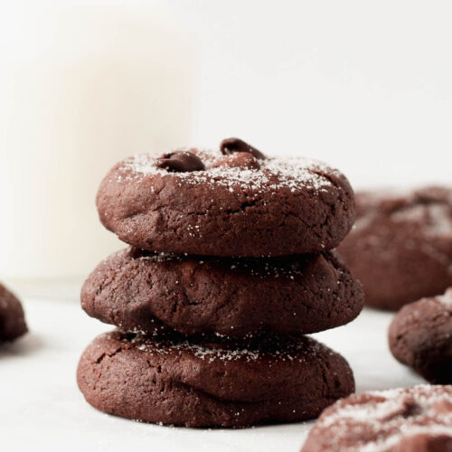 A small stack of double chocolate cookies set on a table next to a glass of milk.