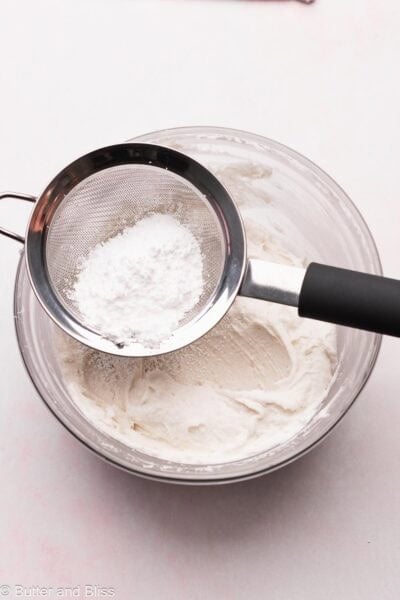Powdered sugar being sifted over a mixing bowl of frosting.