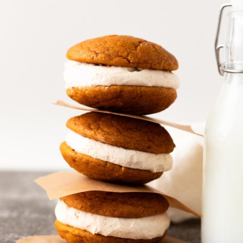 A stack of three pumpkin whoopie pies with cream cheese filling next to a glass of milk.
