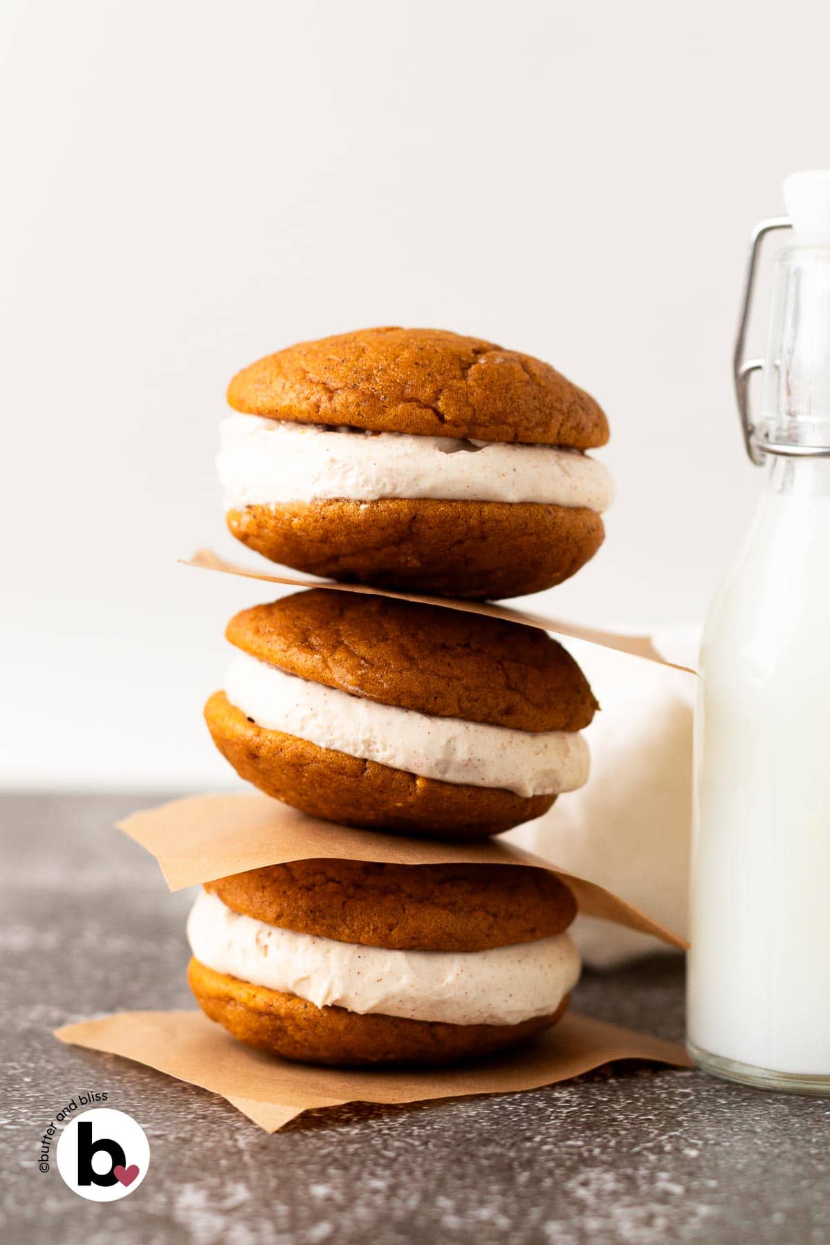 A stack of three pumpkin whoopie pies with cream cheese filling next to a glass of milk.