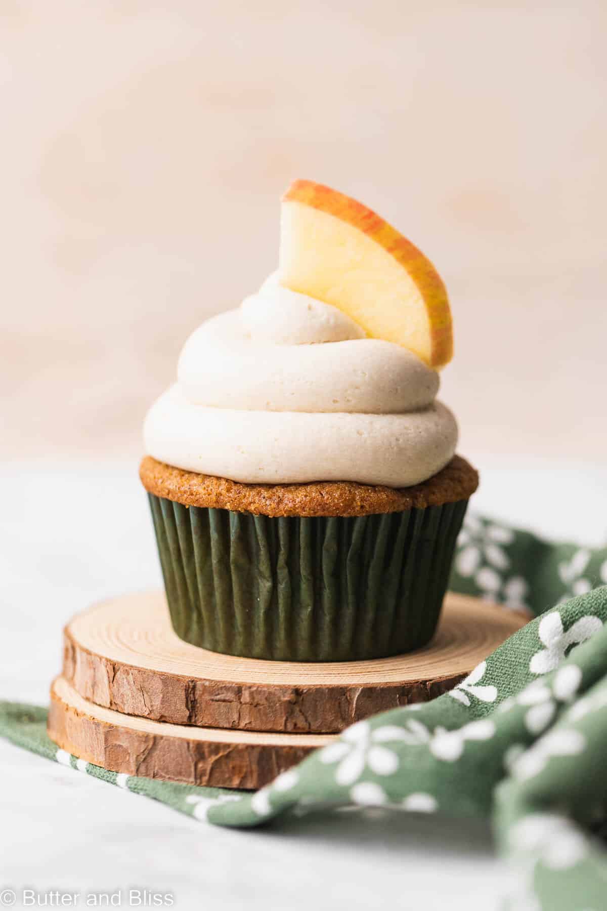 A single gluten free apple cupcake with frosting on a small wood coaster.