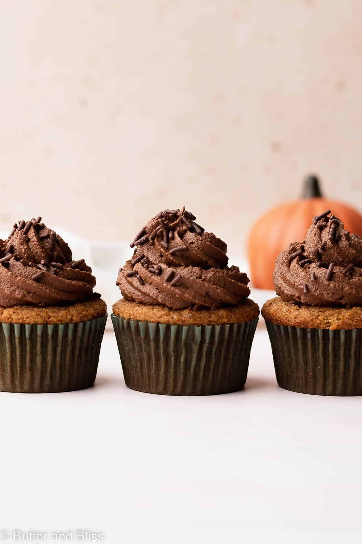 Three pumpkin gluten free cupcakes with chocolate frosting lined up in a row on a table.