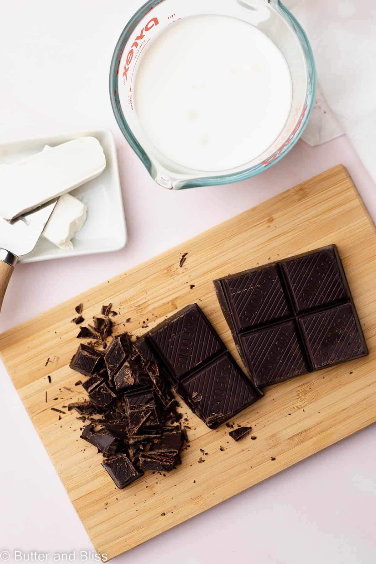 Chocolate bar cut into pieces on a cutting board alongside a measuring cup of cream.