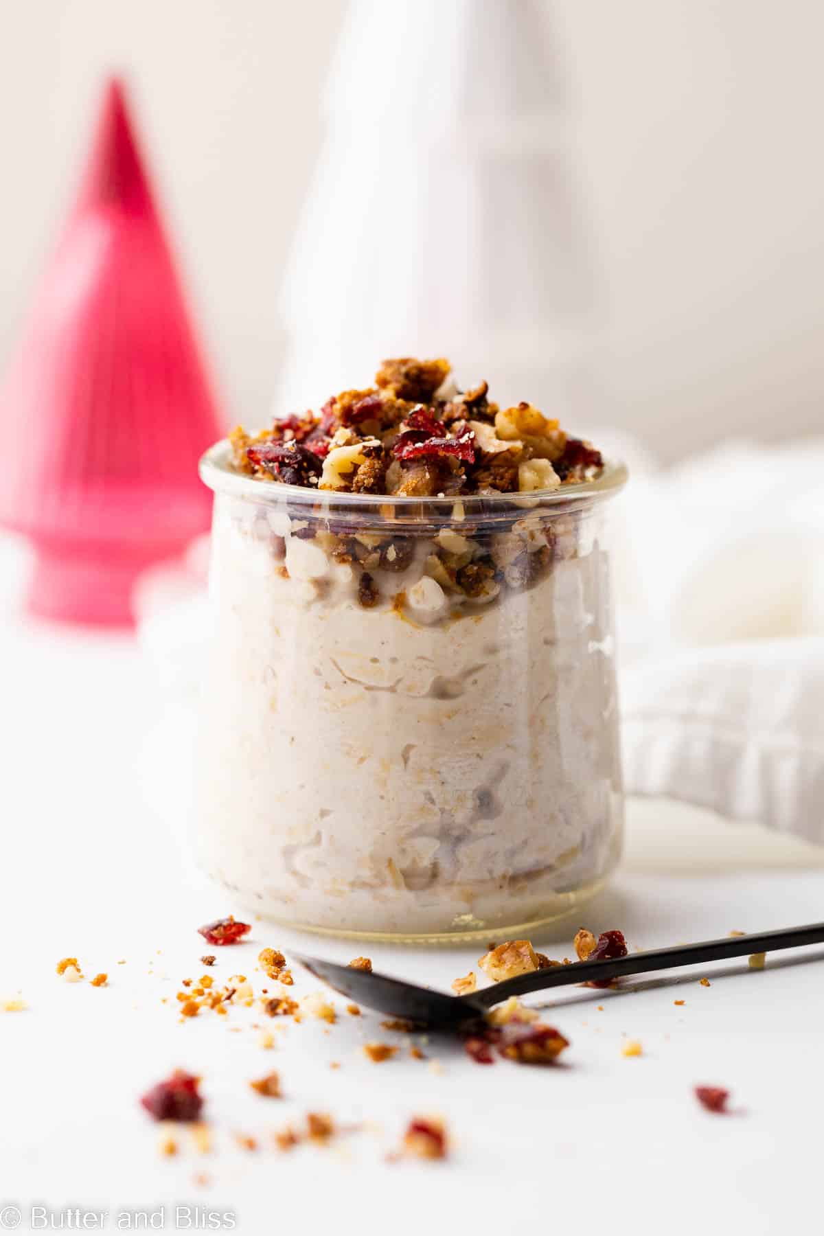 A single serving glass of eggnog overnight oats on a table next to decorative Christmas trees.
