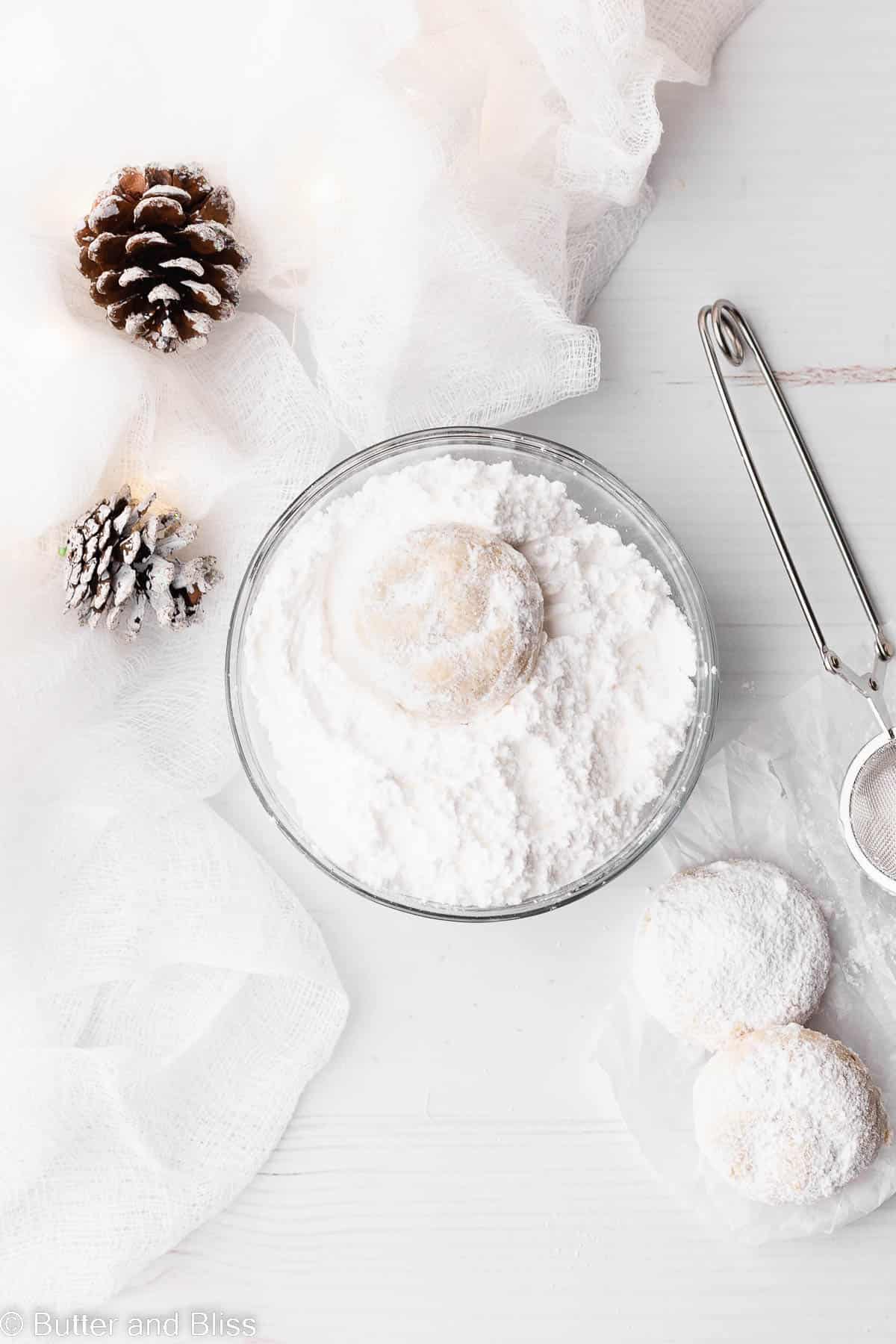 A Christmas cookie being rolled through a bowl of powdered sugar on a white table.
