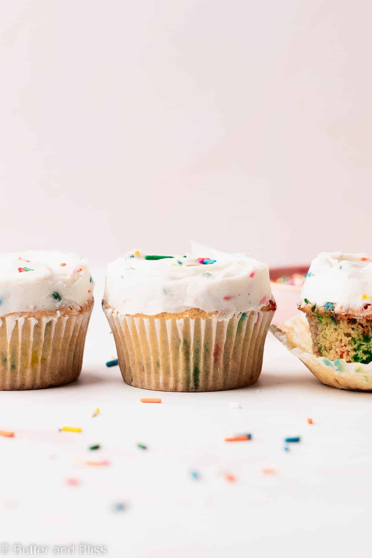 A line up of the cutest vanilla cupcakes with sprinkles lined up on a table.