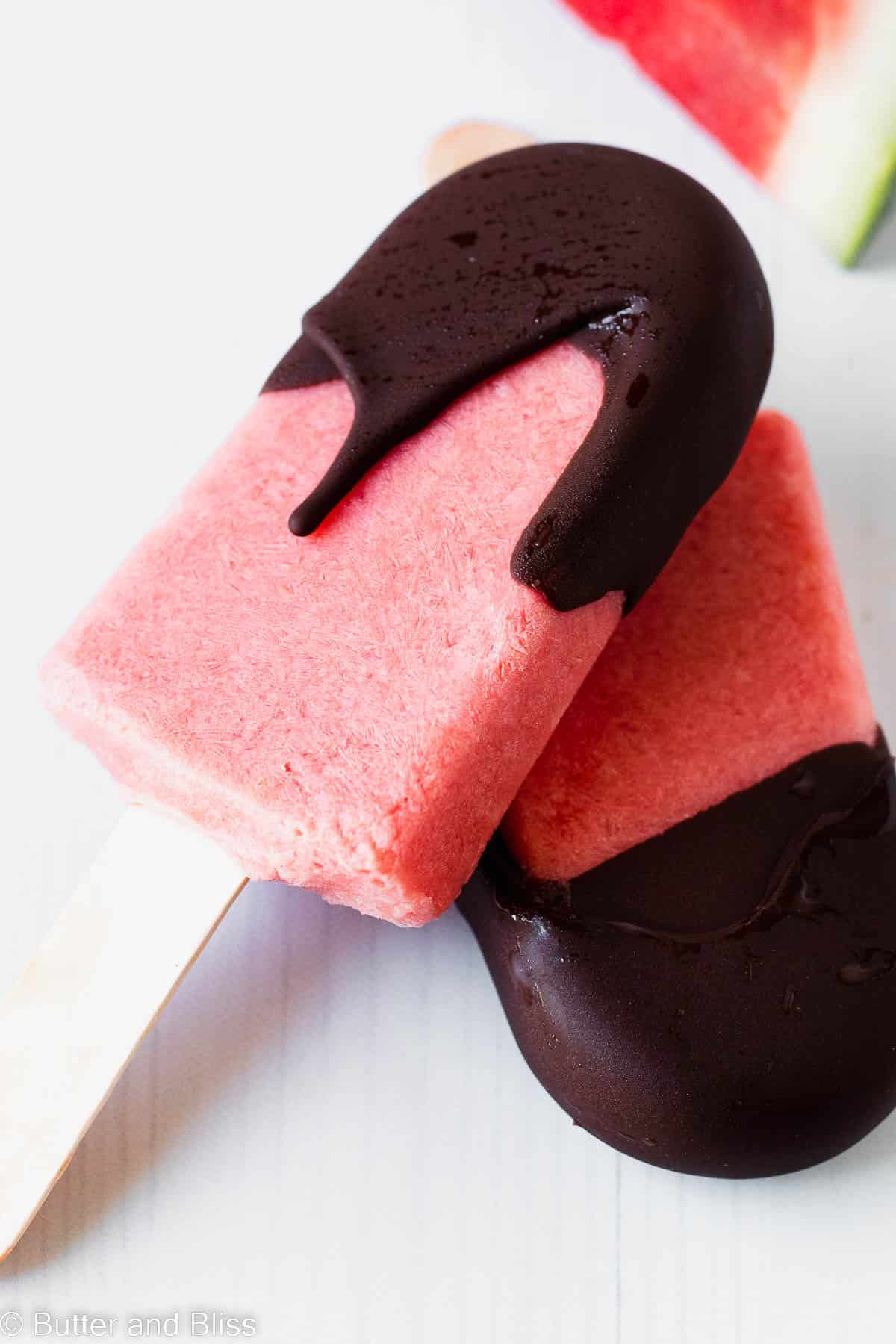 Pretty pink watermelon popsicle with chocolate magic shell next to a watermelon slice.