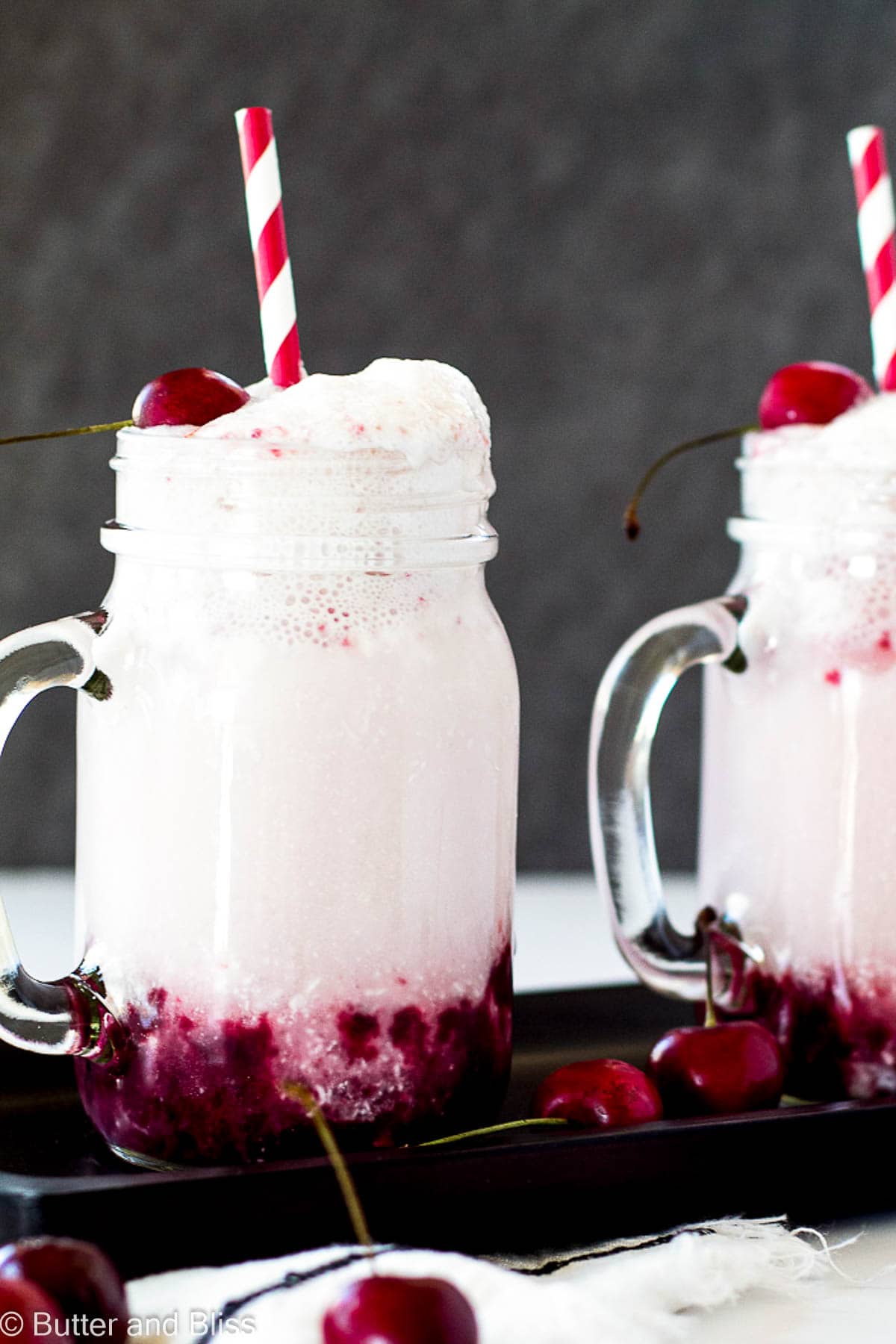 Pretty mugs of black cherry cream soda with red straws and cherry on top.