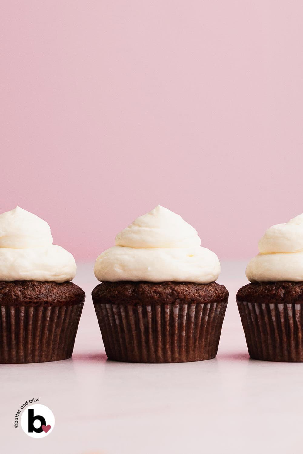 Three chocolate cupcakes with vanilla frosting lined up in a row on a table.