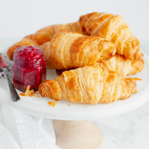 A white serving platter full of buttery homemade croissants with side of jam.