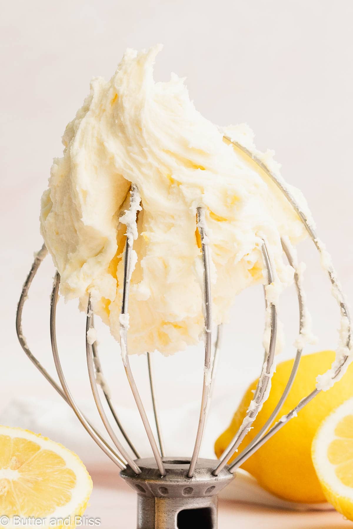 Creamy small batch lemon buttercream frosting on a whisk surrounded by fresh lemon sliced.
