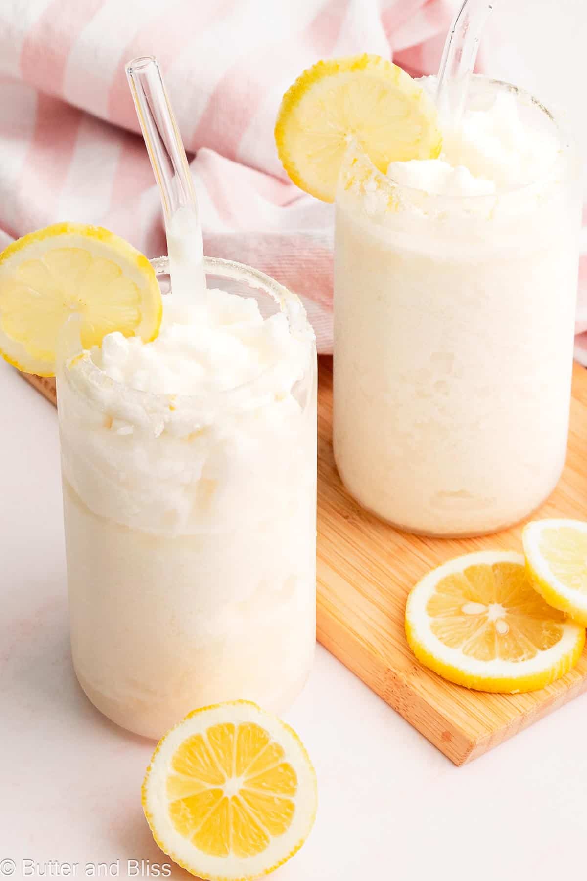 Two glasses of creamy frozen lemonade with lemon wedges and glass straws.