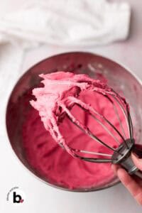 Hand holding a mixing whisk full of bright pink cranberry frosting.