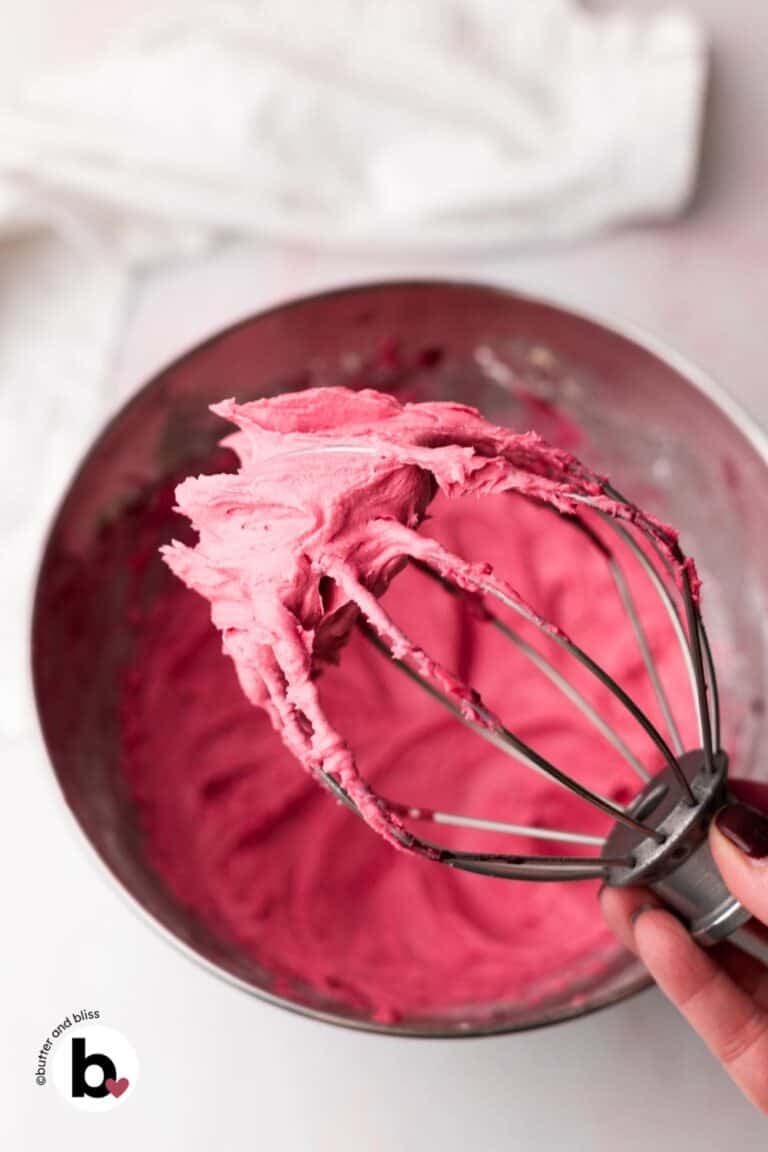 Hand holding a mixing whisk full of bright pink cranberry frosting.