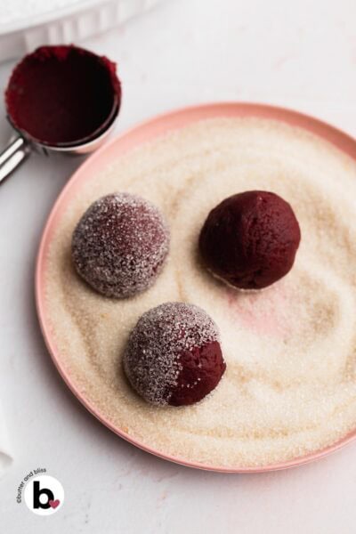 Red tinted cookie dough balls being rolled through granulated sugar.