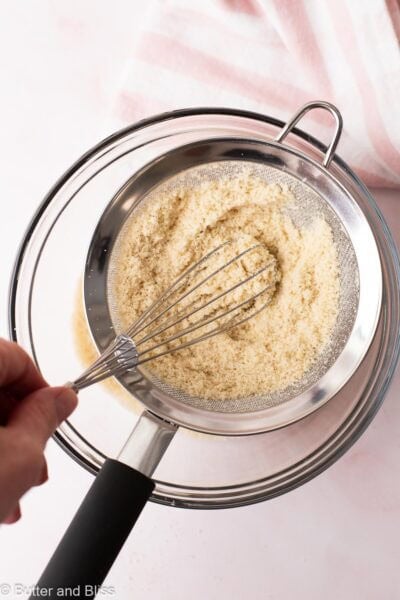 Hand using a whisk to push almond flour through a strainer.