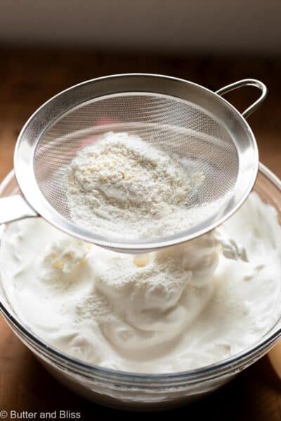 Flour being sifted over beaten egg whites in a mixing bowl.