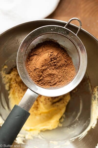 Cocoa powder being sifted into a bowl of whipped butter.
