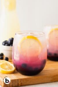 Pretty blueberry lemonade in a glass with lemon wedge and fresh blueberry garnish.