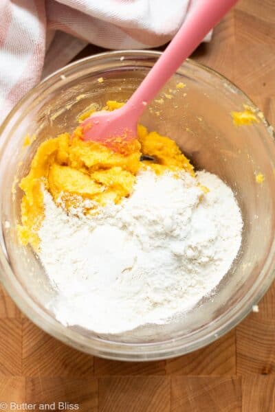 Dry ingredients added to creamed butter in a mixing bowl with a pink spatula.
