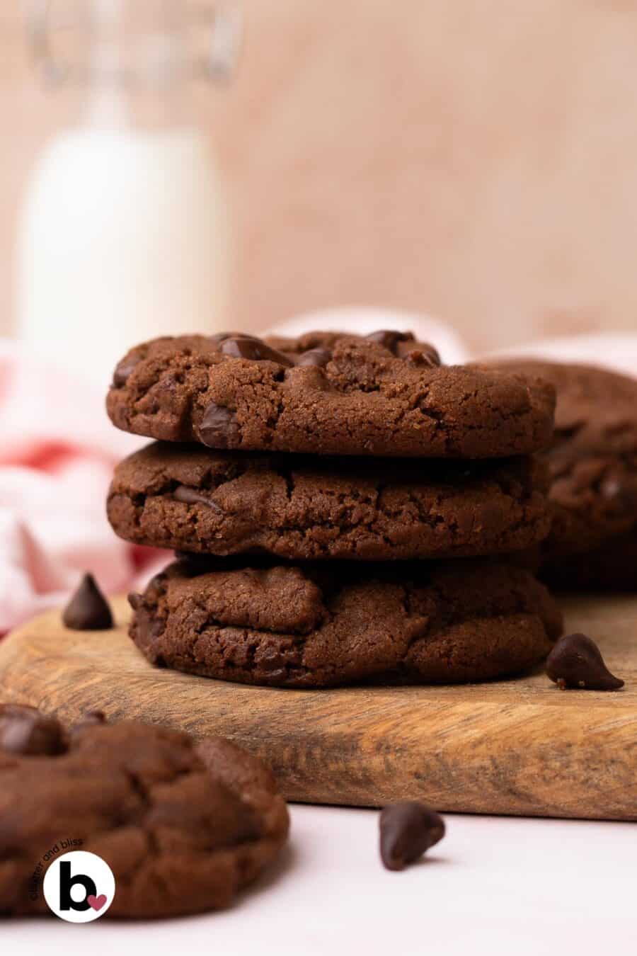 A stack of three double chocolate chip cookies on a wood plate next to a glass of milk.