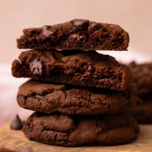 A stack of double chocolate chip cookies on a wood plate with one cut in half to reveal chewy centers.