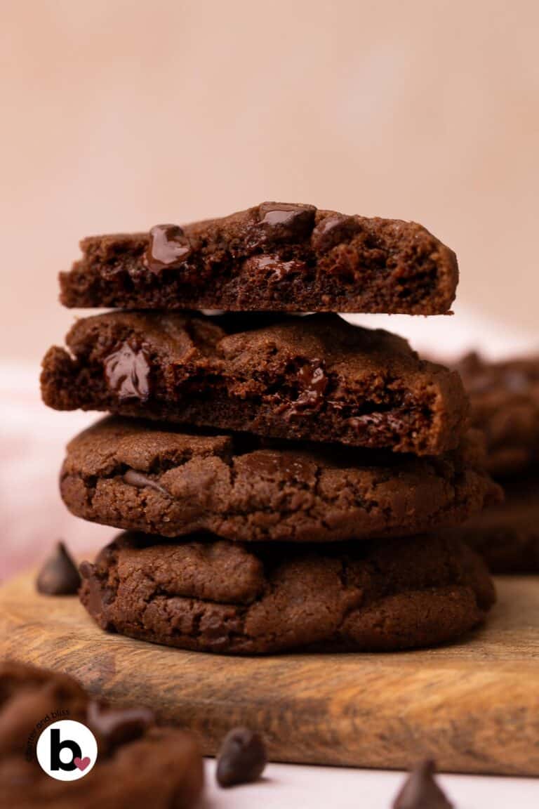 A stack of double chocolate chip cookies on a wood plate with one cut in half to reveal chewy centers.