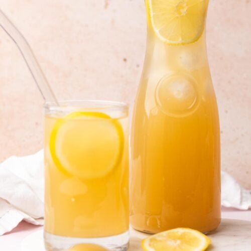 A glass of ginger lemonade next to a pitcher on a marble plate.