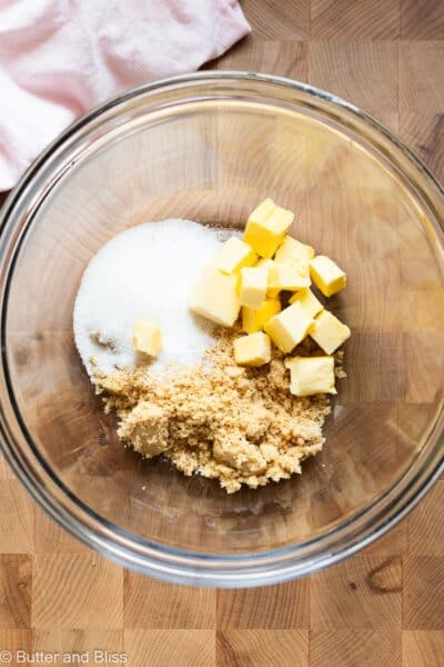 Butter cubes, brown sugar, and white sugar in a mixing bowl on a wood cutting board.