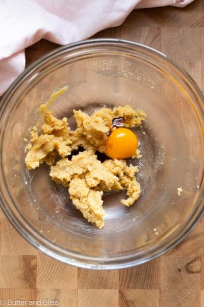 An egg yolk added to a bowl of creamed butter and sugar in a mixing bowl on a wood cutting board.