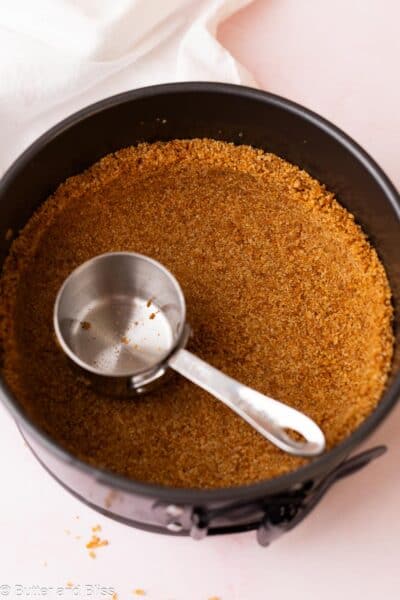 Graham cracker crumbs being pressed into a small pan with a measuring cup.