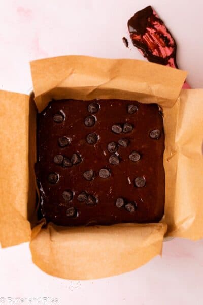 Brownie batter topped with chocolate chips in a small parchment lined square cake pan.