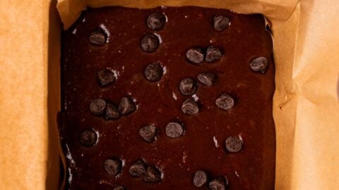 Brownie batter topped with chocolate chips in a small parchment lined square cake pan.