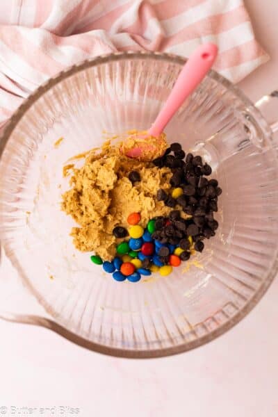 M&M's and chocolate chips added to cookie dough in a clear mixing bowl.