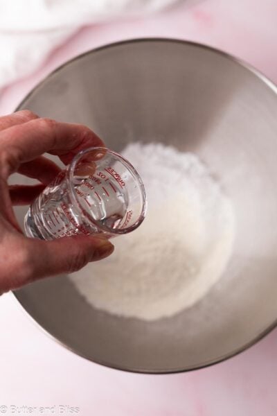 Hand pouring a small amount of water into a mixing bowl of powdered sugar.