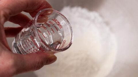 Hand pouring a small amount of water into a mixing bowl of powdered sugar.