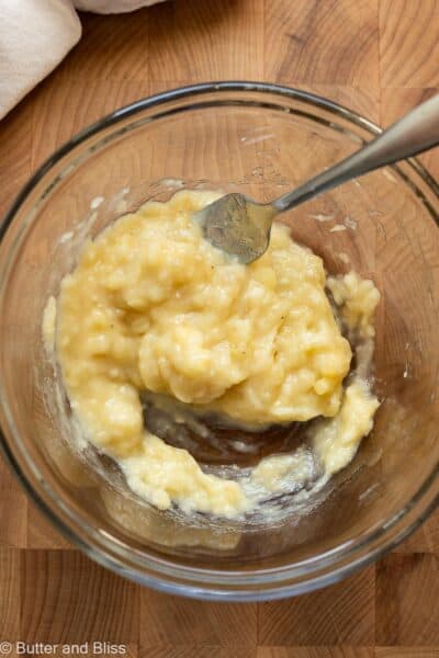 Mashed banana in a bowl with a fork on a wood cutting board.