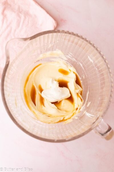 Sour cream and vanilla added to mixed cream cheese in a glass mixing bowl.