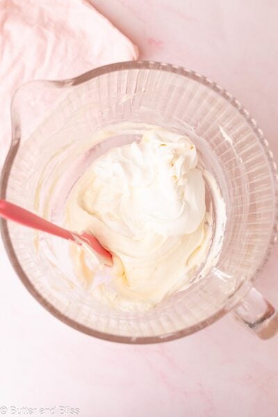 Whipped cream being folded into cream cheese with a pink spatula in a glass mixing bowl.