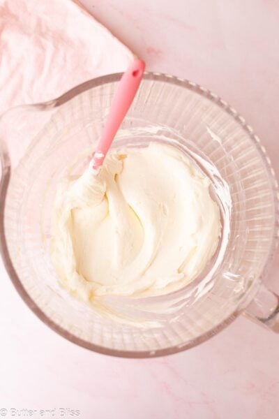 Creamy no-bake cheesecake filling in a glass mixing bowl with a pink spatula.