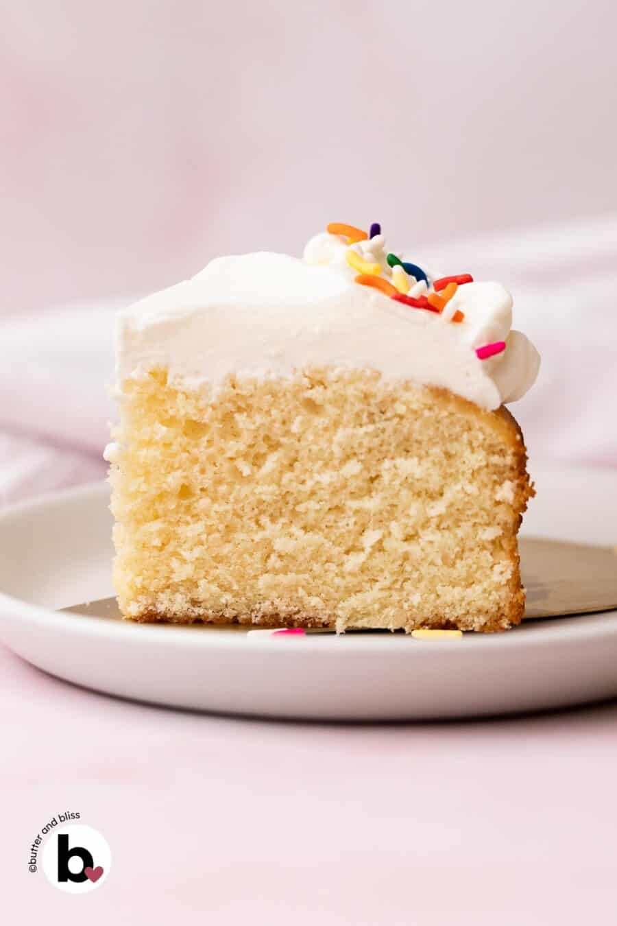 A single slice of white cake with whipped white frosting and colorful sprinkles on a small white plate.