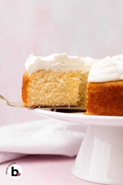 A slice of white cake with white frosting on a cake spatula, cut from the full cake on a white platter.