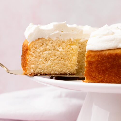 A slice of white cake with white frosting on a cake spatula, cut from the full cake on a white platter.