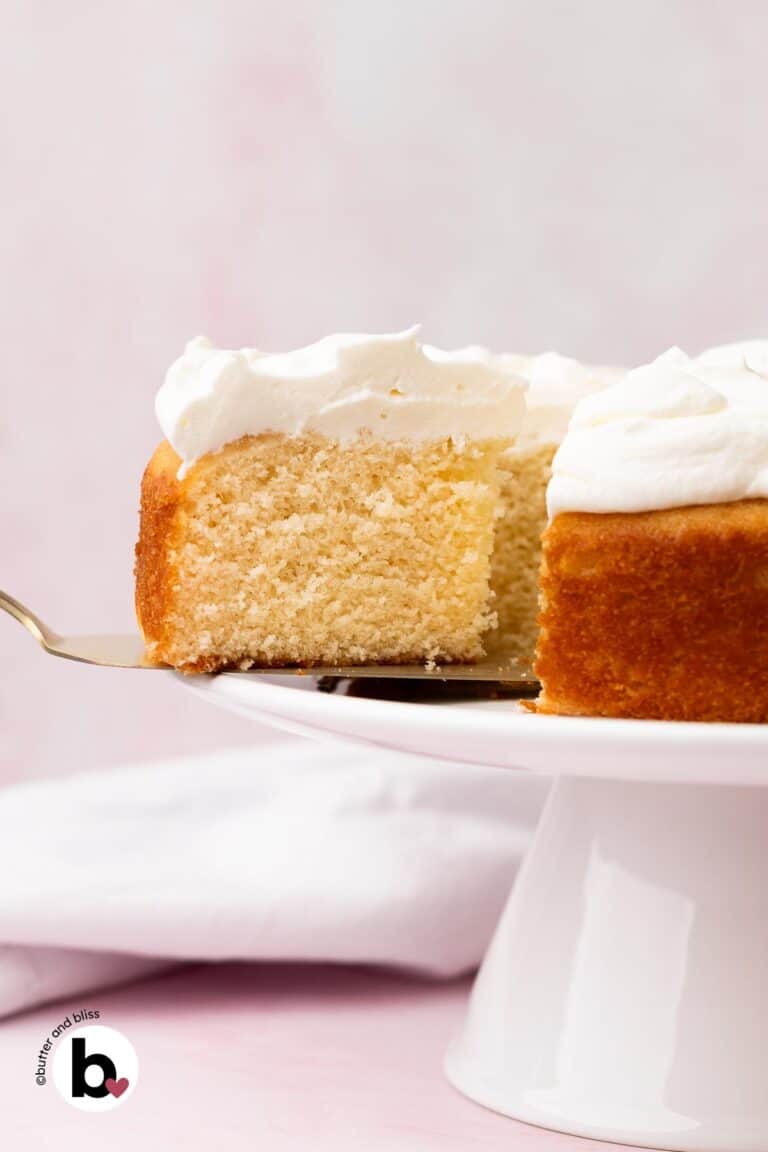 A slice of white cake with white frosting on a cake spatula, cut from the full cake on a white platter.