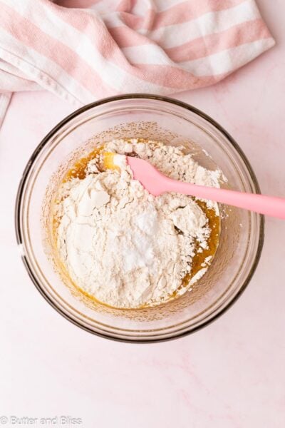 Flour sprinkled over wet cookie bar ingredients in a mixing bowl with a pink spatula.