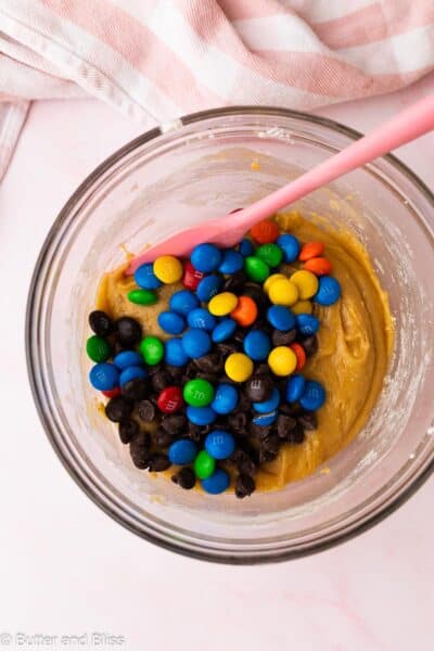 M&M's and chocolate chips added to cookie bar dough in a mixing bowl with a pink spatula.