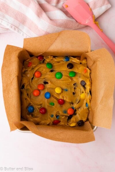 M&M cookie bar dough spread into a square cake pan with a pink spatula.