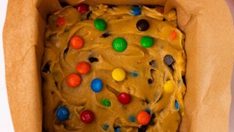 M&M cookie bar dough spread into a square cake pan with a pink spatula.