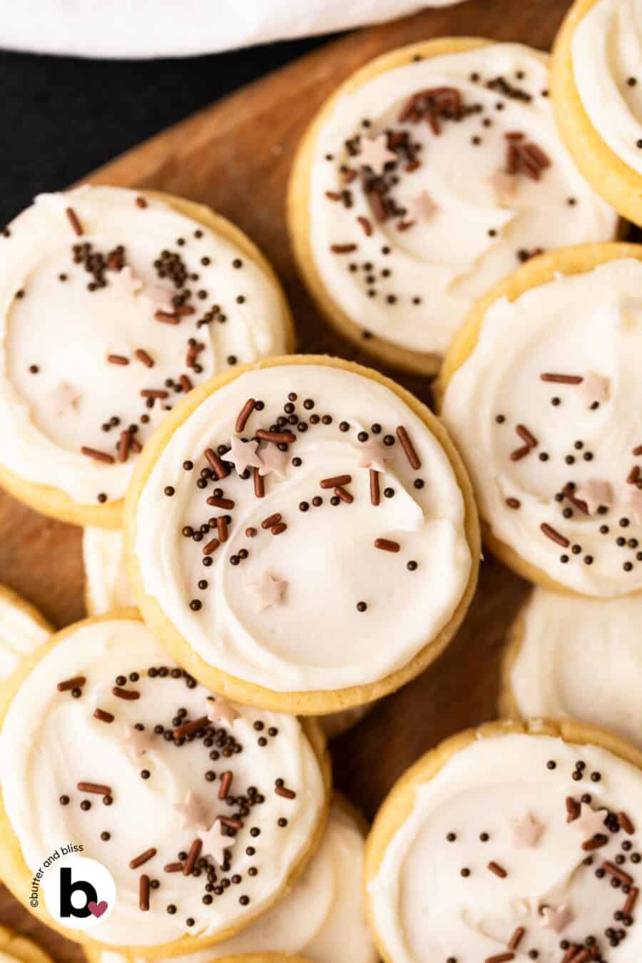 Frosted butter cookies decorated with chocolate sprinkles piled on a wood plate.