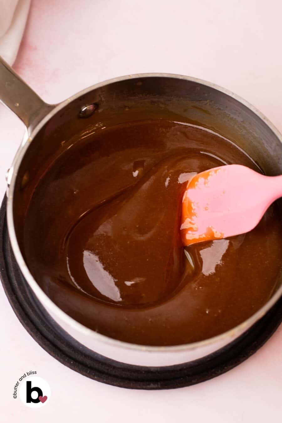 Thick maple syrup caramel sauce in a small saucepan being stirred with a pink spatula.